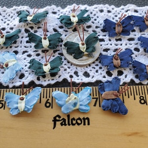May include: Assortment of fabric butterflies in blue, green, and brown colors. The butterflies are arranged on a white lace fabric and a wooden ruler with the word "Falcon" printed on it.
