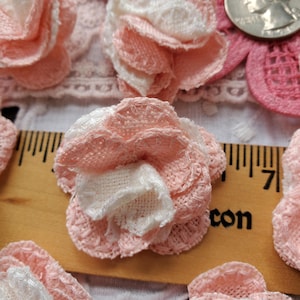 May include: Several layered fabric flower appliques in shades of soft pink and white. The flowers feature a textured, woven appearance with scalloped edges. A wooden ruler is visible underneath, showing the flowers measure approximately 1 inch wide.