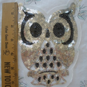 May include: A silver and black sequined owl applique with a white mesh backing. The owl has large, round eyes and a detailed beak.