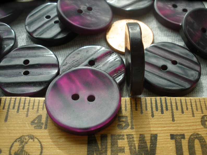 K&ouml;nnte beinhalten: Eine Sammlung von violetten und schwarzen Plastikkn&ouml;pfen mit jeweils zwei L&ouml;chern. Die Kn&ouml;pfe haben ein gestreiftes Muster und eine gl&auml;nzende Oberfl&auml;che.