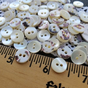 May include: A collection of round, off-white buttons with two holes each. Some buttons have a marbled design in shades of burgundy, brown, and black. A wooden ruler with inch markings is visible, indicating the buttons' size.