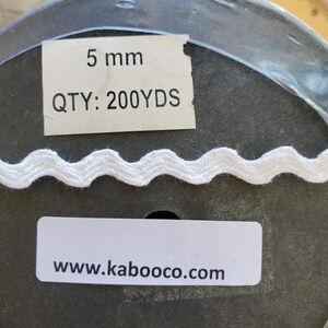 May include: White, wavy trim on a spool. The label reads "5 mm" and "QTY: 200YDS". The website address www.kaboooco.com is also visible. This trim is suitable for various sewing and crafting projects.