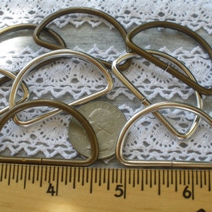 May include: A close-up of a group of metal D-rings in two finishes: antique brass and silver. The rings are arranged on a white lace fabric background with a ruler for scale. The rings measure approximately 1 inch in diameter.
