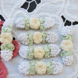May include: A set of six handmade fabric flower embellishments. Each embellishment features a white, fluffy center, a pale yellow satin flower, and green satin leaves.