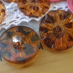 May include: A set of four amber glass buttons with a swirl design. The buttons have a shiny finish and a raised design.