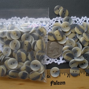 May include: A collection of round buttons with two holes, in a mix of gray and tan. Some buttons are in a clear plastic bag, others are loose. A quarter is next to the buttons for size comparison. The buttons are on a wooden surface.