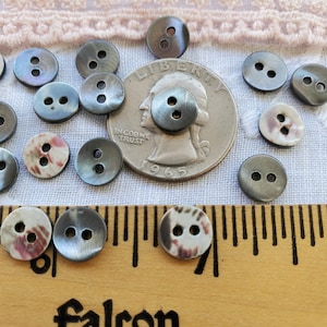 May include: A collection of 18 two-hole buttons in various colors and patterns. The buttons are arranged on a white fabric surface with a US quarter for scale. Some buttons are black with a pearlescent sheen, while others are white with a pink and brown marbled pattern.