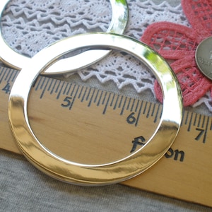 May include: Two silver metal rings, one larger than the other, are placed on top of a wooden ruler with inch markings. The rings are shiny and have a smooth, polished finish.