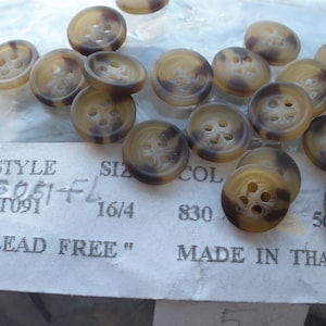 May include: A collection of round, tortoiseshell-patterned buttons with four holes. The buttons are a mix of tan and brown, with a marbled effect. The text "LEAD FREE" and "MADE IN THAILAND" are visible on a label.