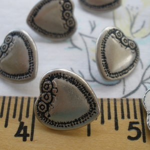 May include: Silver heart-shaped buttons with a decorative border and three small circles in the center. The buttons are arranged on a white surface with a ruler.