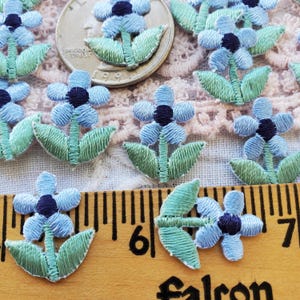 May include: Embroidered blue flower appliques with dark blue centers and green leaves. The appliques are arranged on a pink lace surface with a ruler and a quarter for scale. These floral appliques are ideal for crafting and sewing projects.