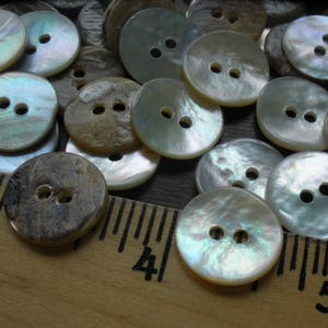 May include: A collection of iridescent white and brown mother of pearl buttons with two holes each. The buttons are arranged in a random pattern on a wooden surface with a ruler.