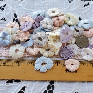 May include: A collection of small, embroidered flower appliques in pastel colors, including blue, pink, lavender, and cream. The flowers are arranged on a white lace background with a wooden ruler for scale. A US quarter is also present.