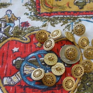May include: A collection of 12 gold-toned metal buttons with intricate designs. The buttons are arranged on a colorful fabric background.