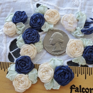 May include: A collection of 12 handmade fabric flowers in shades of ivory and navy blue. The flowers are arranged on a white lace fabric background.  The flowers are approximately the size of a US quarter.