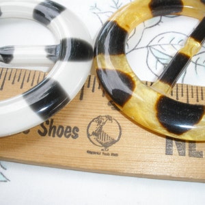 May include: Two vintage belt buckles. One is white with black stripes, the other is a tortoiseshell pattern of amber and black. Both are round with a rectangular bar for the belt.
