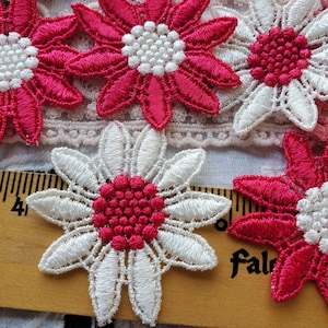 May include: A collection of embroidered flower patches in bright pink and white, featuring detailed stitching and textured centers. A wooden ruler is visible underneath, showing the flowers measure approximately 1.5 inches across.