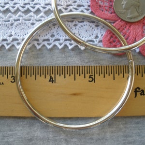 May include: Two silver hoop earrings, approximately 3 inches in diameter, are displayed on a wooden ruler. The ruler is marked with black lines and numbers. A quarter and a pink flower are in the background.