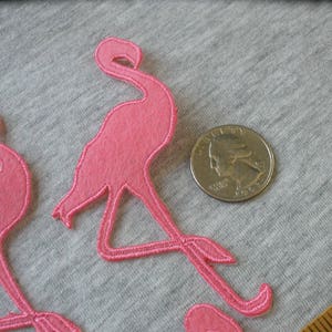 May include: Pink flamingo applique patches with embroidered details. The patches are next to a quarter for size reference. The patches are made of felt and have a stitched outline. The flamingo patches are a fun craft supply.