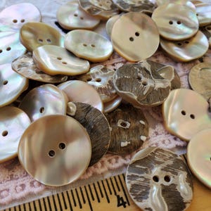 May include: A collection of assorted buttons in various shapes and sizes. The buttons are primarily cream and brown, with some featuring a pearlescent sheen. Some buttons have two holes, while others have four. The buttons are scattered on a light pink surface.