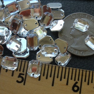 May include: A close-up of a pile of clear, faceted, octagon-shaped rhinestones with silver backing. The rhinestones are arranged on a wooden ruler with inch markings. A US quarter is included for size reference.