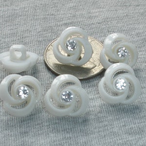 May include: Six white plastic buttons with a swirl design and a clear rhinestone in the center. The buttons are approximately the size of a US quarter.