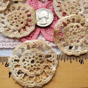 May include: Close-up of several beige and cream-colored lace doilies with a quarter coin for scale. The doilies have a circular design with intricate patterns. A pink lace flower and a wooden ruler are also visible.