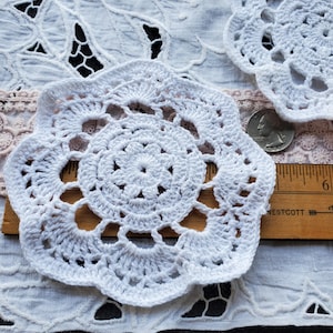 May include: Close-up of two white, handmade crochet doilies with a floral design. The doilies are placed on a white lace surface with a wooden ruler and a quarter for size comparison. The doilies have a scalloped edge.