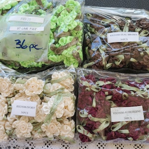 May include: Five bags of fabric flower embellishments. The bags are labeled with the color and quantity of the flowers. The colors are lime green, brown, ivory, and burgundy. The bags are labeled "RB 20 97 Lime Fizz 36pc", "RB20SM-SEAL BROWN 36PCS", "RB20SM-IVORY 36PCS", and "RB20SM-AUBERGINE 36PCS".