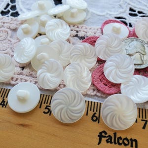 May include: A collection of round, white buttons with a swirling, textured pattern on the surface. Each button features a central shank on the back. A wooden ruler is visible at the bottom, showing the buttons are approximately 0.5 inches wide.