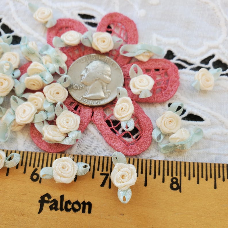 May include: Close-up of small, cream-colored satin roses with light blue ribbon accents. The roses are scattered around a quarter coin and a pink lace applique. A wooden ruler with the word "Falcon" is in the foreground.