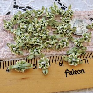 May include: A collection of small, green ribbon flowers with white pearl centers. The flowers are arranged on a pink lace surface with a wooden ruler and a quarter for scale. The ruler has the word "Falcon" printed on it.
