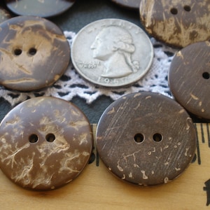 May include: Close-up of several large, round, brown coconut buttons with two holes each. A silver quarter coin is visible, along with a ruler. The buttons have a natural, textured appearance.