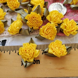 May include: A collection of small yellow ribbon roses with olive green leaves, each crafted from folded fabric. Two of these floral embellishments are positioned directly on a wooden ruler, showing they measure approximately 0.75 inches across.