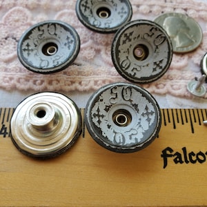May include: Close-up of several vintage metal buttons with a distressed, aged appearance. The buttons are primarily gray with black edging and feature the number "50" along with decorative cross-like symbols. A ruler and a coin are also visible.