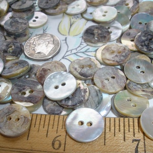 May include: A large collection of round, white, and brown mother-of-pearl buttons with two holes each. The buttons are arranged in a random pattern on a wooden surface with a ruler.