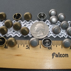 May include: A collection of 17 metal buttons in two colors: antique brass and silver. The buttons are round and have a raised, textured design. They are arranged on a white lace doily and a wooden ruler.