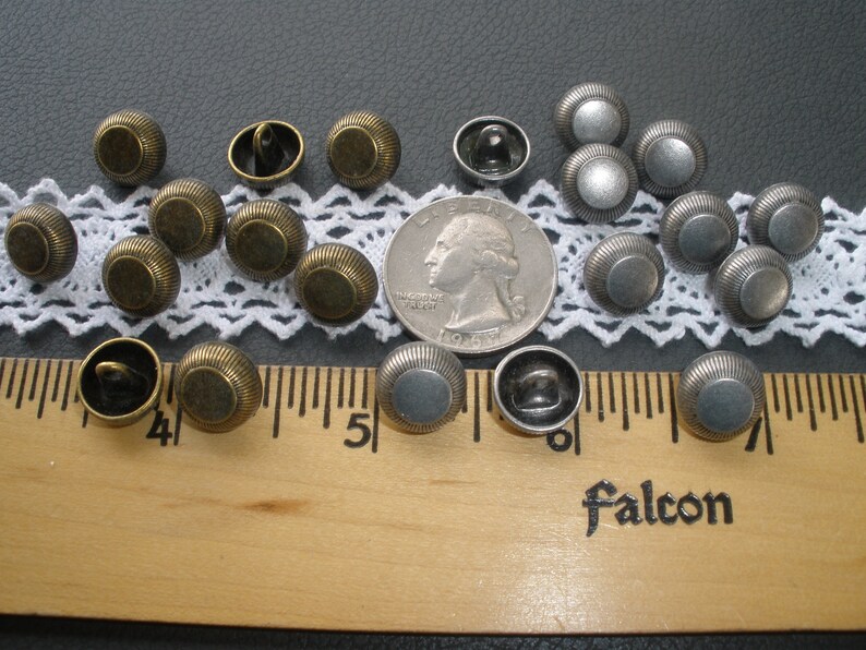May include: A collection of 17 metal buttons in two colors: antique brass and silver. The buttons are round and have a raised, textured design. They are arranged on a white lace doily and a wooden ruler.