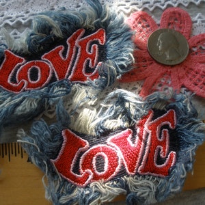 May include: Two denim patches with frayed edges, each featuring the word "LOVE" embroidered in red with white outlines. The patches are on a white lace background.