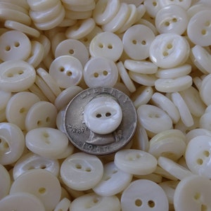 May include: A collection of off-white, pearlescent buttons with two holes each. The buttons are round and of varying sizes, with a U.S. quarter for size comparison. The buttons are scattered, creating a textured surface.