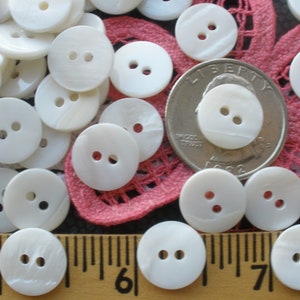 May include: A collection of white, round, two-hole shell buttons. The buttons are arranged on a pink and white fabric background with a US quarter for scale.