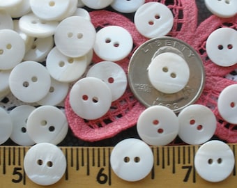 Könnte beinhalten: Eine Sammlung weißer, runder, zweilöchriger Muschelknäpfe. Die Knäpfe sind auf einem rosa-weißen Stoffhintergrund mit einem US-amerikanischen Vierteldollar zur Skalierung angeordnet.