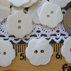May include: A collection of white, flower-shaped buttons with two holes each. The buttons are arranged on a white lace doily and a wooden ruler. A US quarter is also present for size comparison.