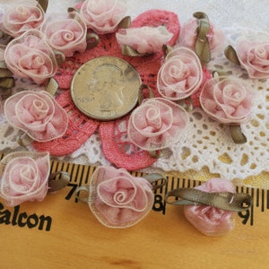 May include: A collection of pink and white fabric rosettes with brown ribbon accents. The rosettes are arranged on a white lace doily and a wooden ruler.