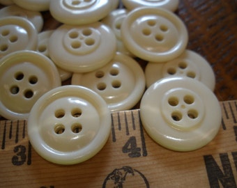 May include: A collection of off-white plastic buttons with four holes each. The buttons are arranged in a semi-circle on a wooden surface with a ruler.