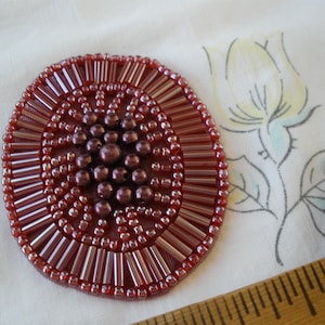 May include: A round, burgundy-colored beaded embellishment with a central cluster of smaller beads. The embellishment is made of glass beads and has a raised, textured design.