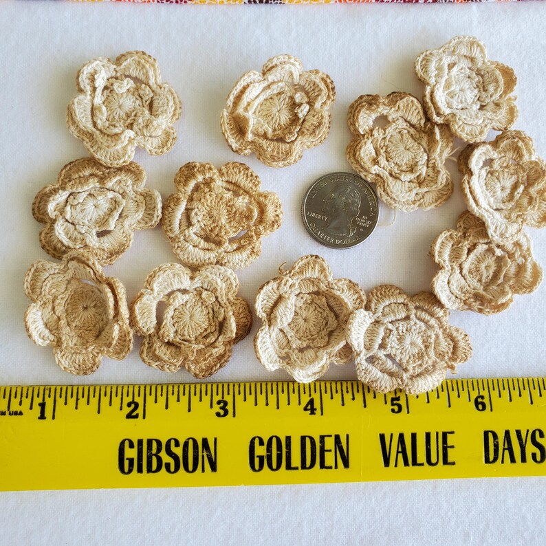 May include: A collection of crocheted flower appliques in shades of beige and tan. The flowers are arranged on a white surface with a yellow ruler and a US quarter for size reference. The flowers have a detailed, textured appearance.