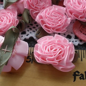 May include: A close-up of pink fabric flowers with green ribbon accents. The flowers are arranged on a white crocheted surface with a wooden ruler in the foreground.