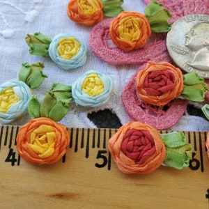May include: Assortment of handmade fabric flowers in various colors, including blue, yellow, orange, and red. The flowers are made from ribbon and have a layered, ruffled appearance. The flowers are arranged on a white fabric background with a ruler and a US quarter for scale.