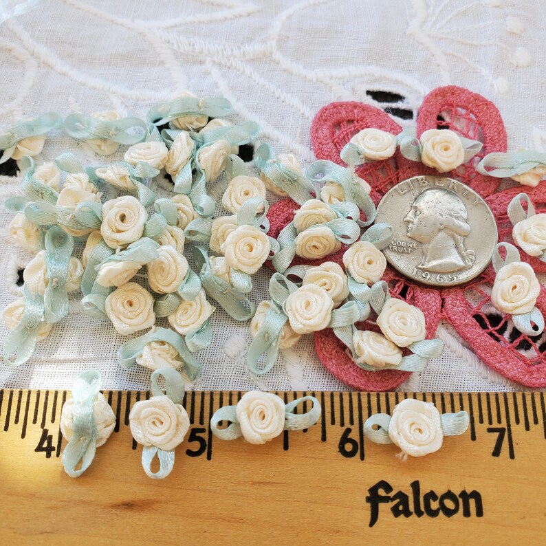 May include: A collection of small, cream-colored satin ribbon roses with light blue ribbon accents. The roses are scattered on a white lace surface, with a wooden ruler and a US quarter for size reference. The word "Falcon" is printed on the ruler.